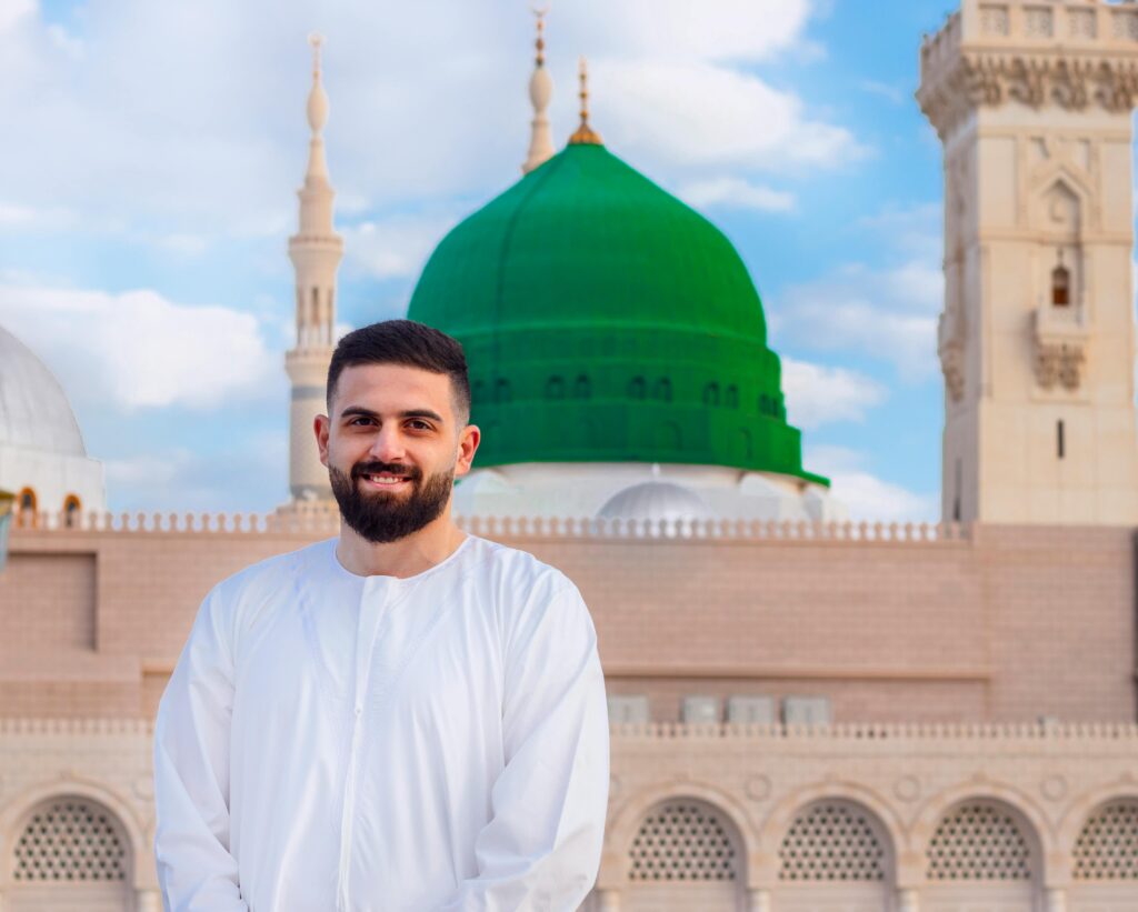 A bearded man smiling in front of the iconic green dome of Al-Masjid an-Nabawi, Medina.