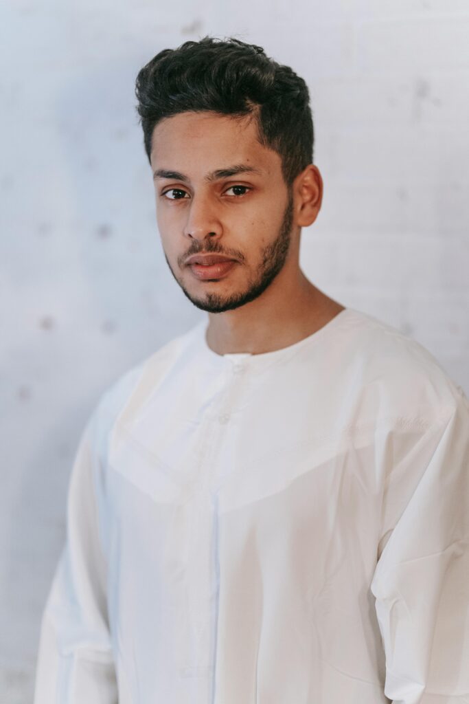 A serious man wearing a white thobe, standing indoors against a light background.